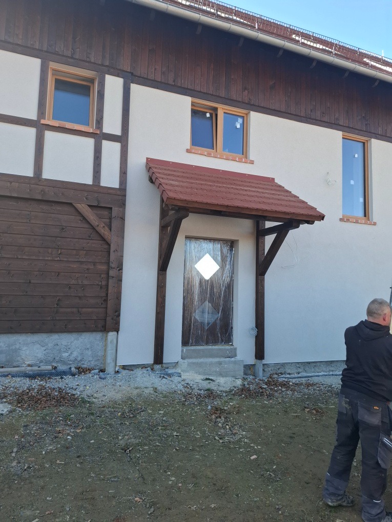 Budynek po ociepleniu elewacji z drewnianymi oknami i zadaszeniem nad wejściem, widoczne ślady prac wykończeniowych.