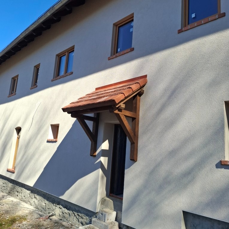 Elewacja budynku z nową strukturą, drewniane obramowania okien oraz zadaszenie nad wejściem z drewnianą konstrukcją i dachówką.