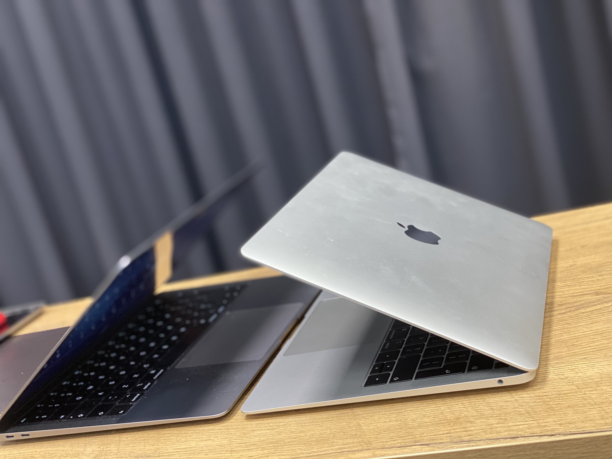 Dwa laptopy MacBook na drewnianym biurku, jeden otwarty, drugi tylko częściowo, z widocznym logo Apple na obudowie.