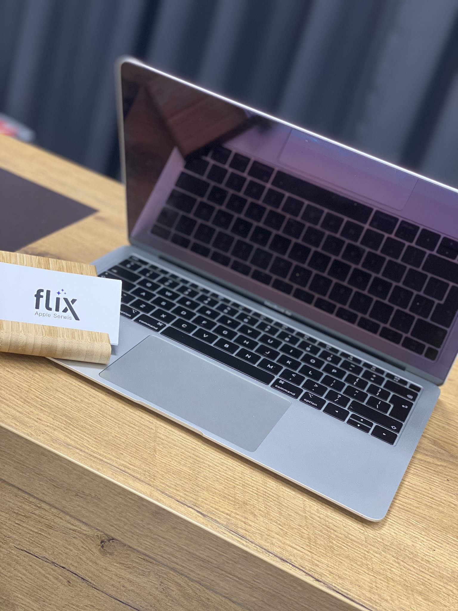 Otwarty laptop Apple na drewnianym biurku, obok wizytówka serwisu Apple w bambusowym stojaku, widoczne odbicie klawiatury w ekranie.