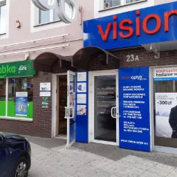 Fasada budynku z szyldem 'vision optyk' i wejściem do salonu optycznego, obok sklep Żabka Cafe, zaparkowany samochód na pierwszym planie.