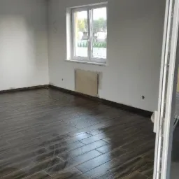 Pokój z ciemnymi, drewnopodobnymi płytkami na podłodze, grzejnik zabezpieczony kartonem, widok przez okno na domy w oddali.