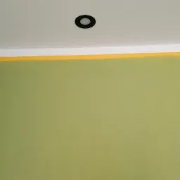 Ściana pomalowana na oliwkowy kolor z widoczną żółtą taśmą malarską przy suficie i pojedynczym, okrągłym punktem oświetleniowym wpuszczonym w sufit.