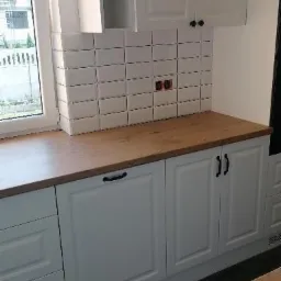 Fragment białej zabudowy kuchennej z drewnianym blatem i białymi płytkami ułożonymi w kwadraty na ścianie, widoczne gniazdka elektryczne, czarne uchwyty szafek.