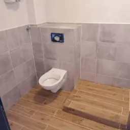 Wykończona łazienka z szarymi płytkami ściennymi i podłogą imitującą drewno, widoczny podwieszany sedes i fragment instalacji sanitarnej.