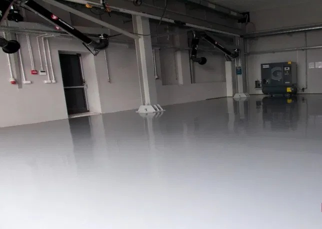 Błyszcząca, szara podłoga przemysłowa w hali z widocznymi instalacjami wentylacyjnymi pod sufitem i kompresorem powietrza w tle.