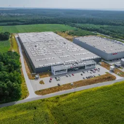 Zdjęcie z lotu ptaka przedstawia dwa duże budynki magazynowe z parkingiem, otoczone zielenią pól i lasów, z turbinami wiatrowymi widocznymi w oddali.