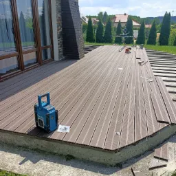 Montaż kompozytowej deski tarasowej na betonowej wylewce, widoczne narzędzia, radio budowlane Makita i elementy mocujące, w tle dom z ceglanym kominem i zielony ogród.