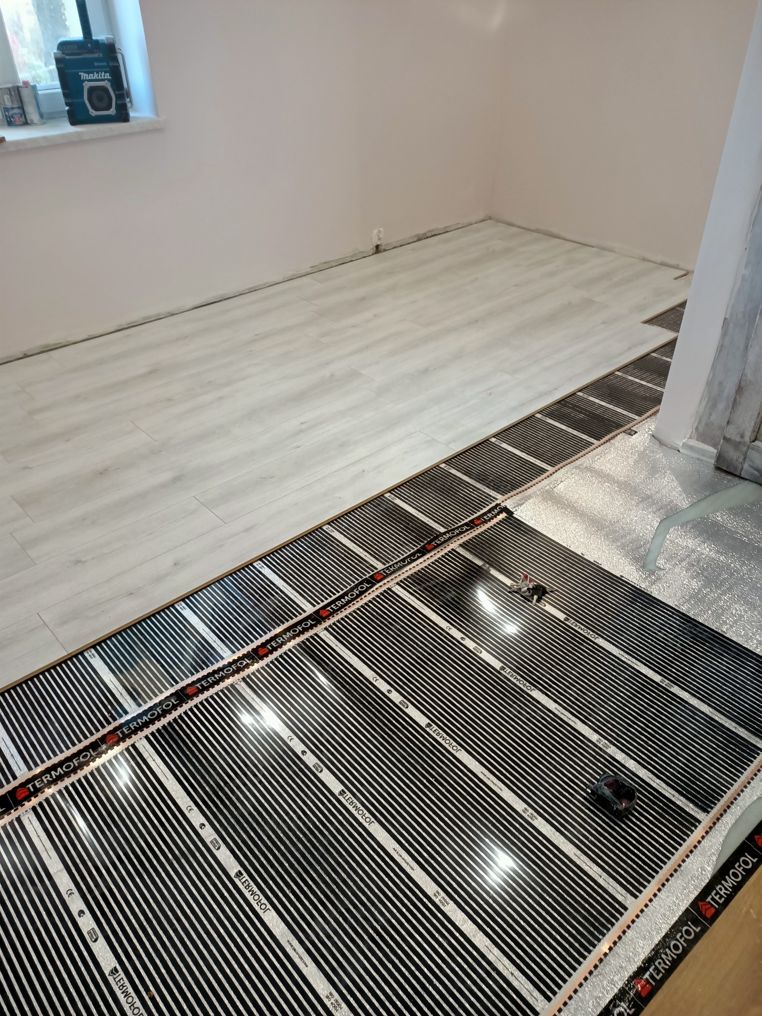 Montaż folii grzewczej Termofol na podłodze w trakcie remontu, widoczny fragment paneli podłogowych i narzędzie do docinania folii.