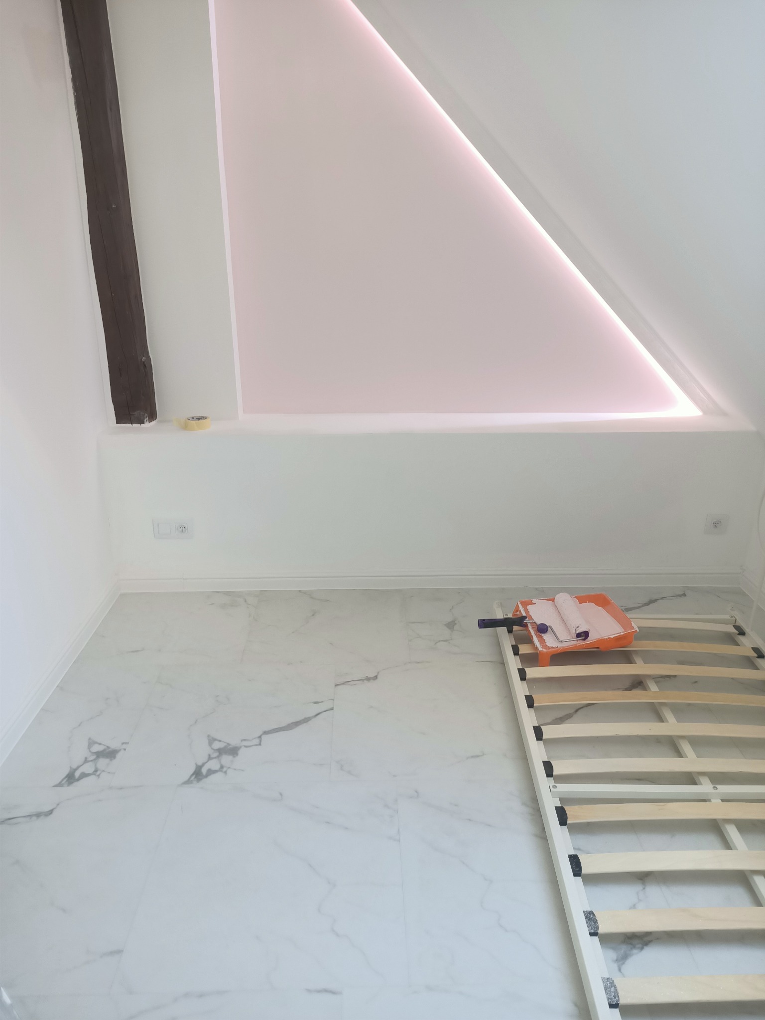 Poddasze po malowaniu, z widoczną drewnianą belką, białymi ścianami, marmurową podłogą, napinanym sufitem z oświetleniem LED, wałkiem i kuwetą z farbą oraz rozłożonym stelażem łóżka.