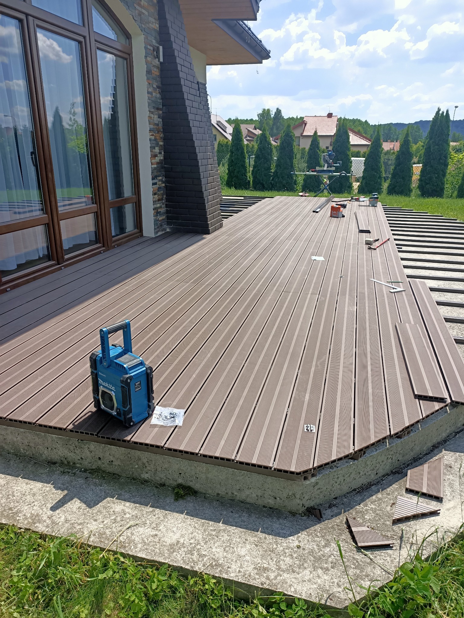 Montaż kompozytowej deski tarasowej na betonowej wylewce, widoczne narzędzia, radio budowlane Makita i elementy mocujące, w tle dom z ceglanym kominem i zielony ogród.