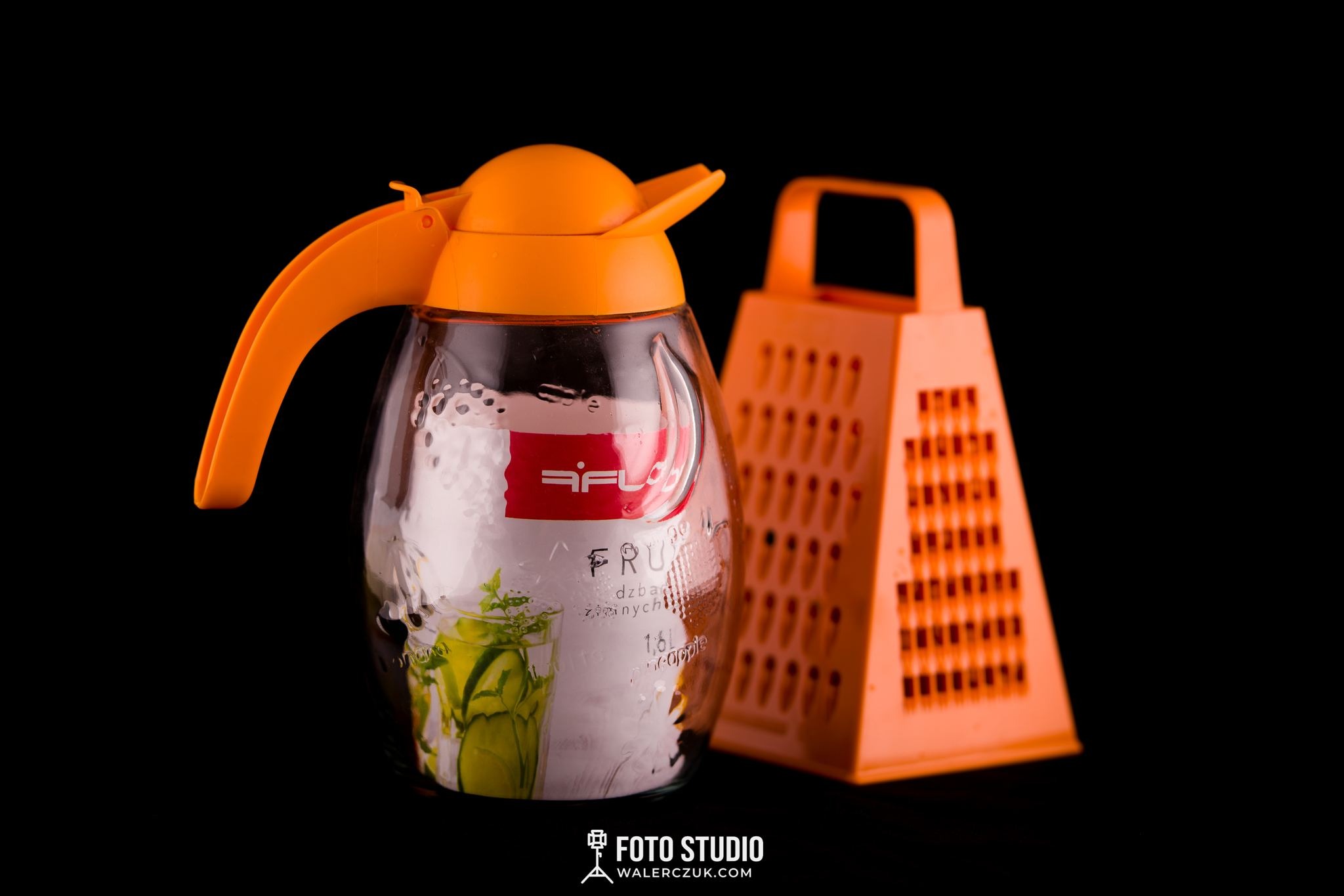 Szklany dzbanek z pomarańczową pokrywką i uchwytem, wypełniony wodą z plasterkami ogórka i ziołami, obok pomarańczowa tarka na czarnym tle, studyjne oświetlenie.