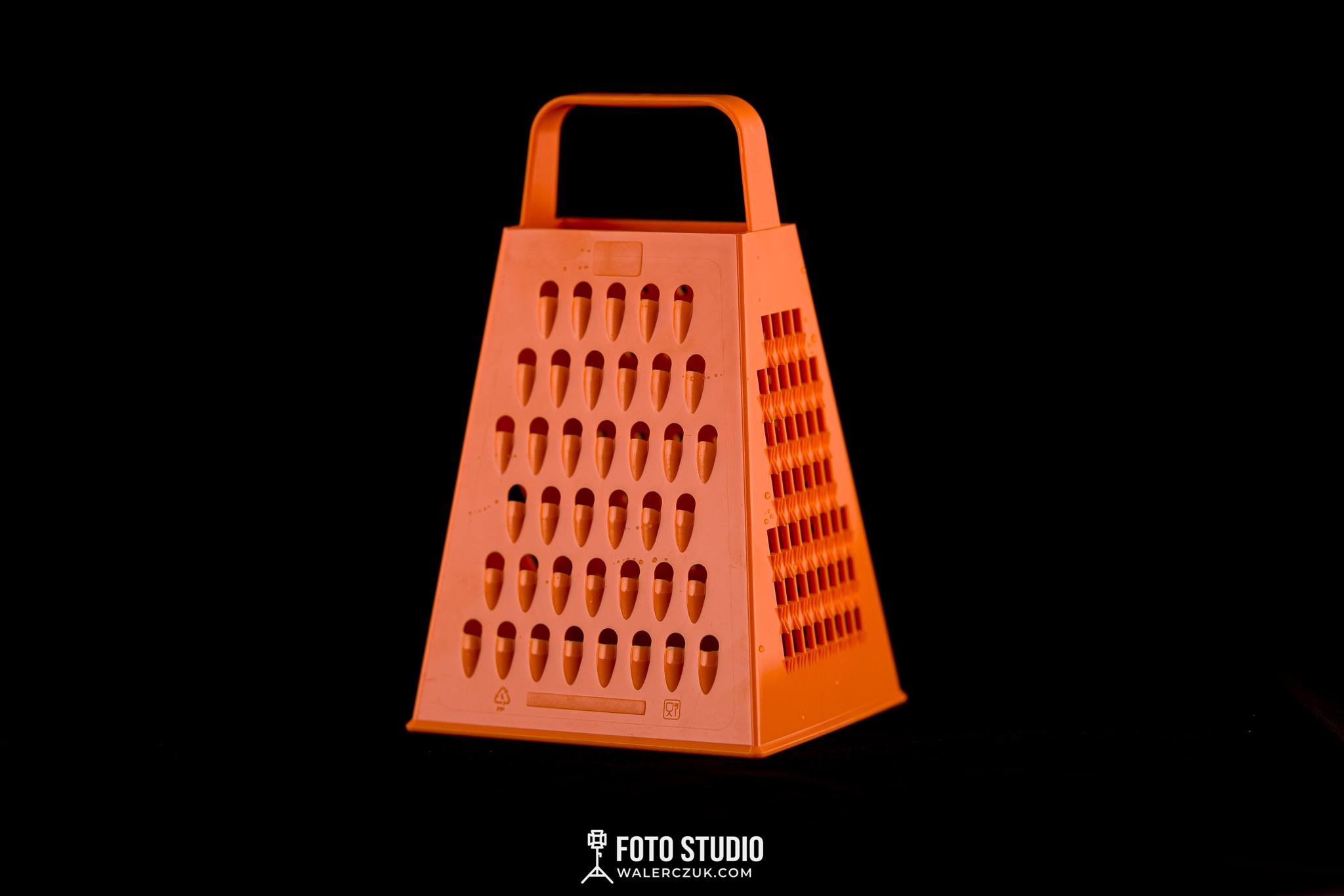 Pomarańczowa, plastikowa tarka o czterech ścianach na czarnym tle, oświetlona studyjnym światłem.