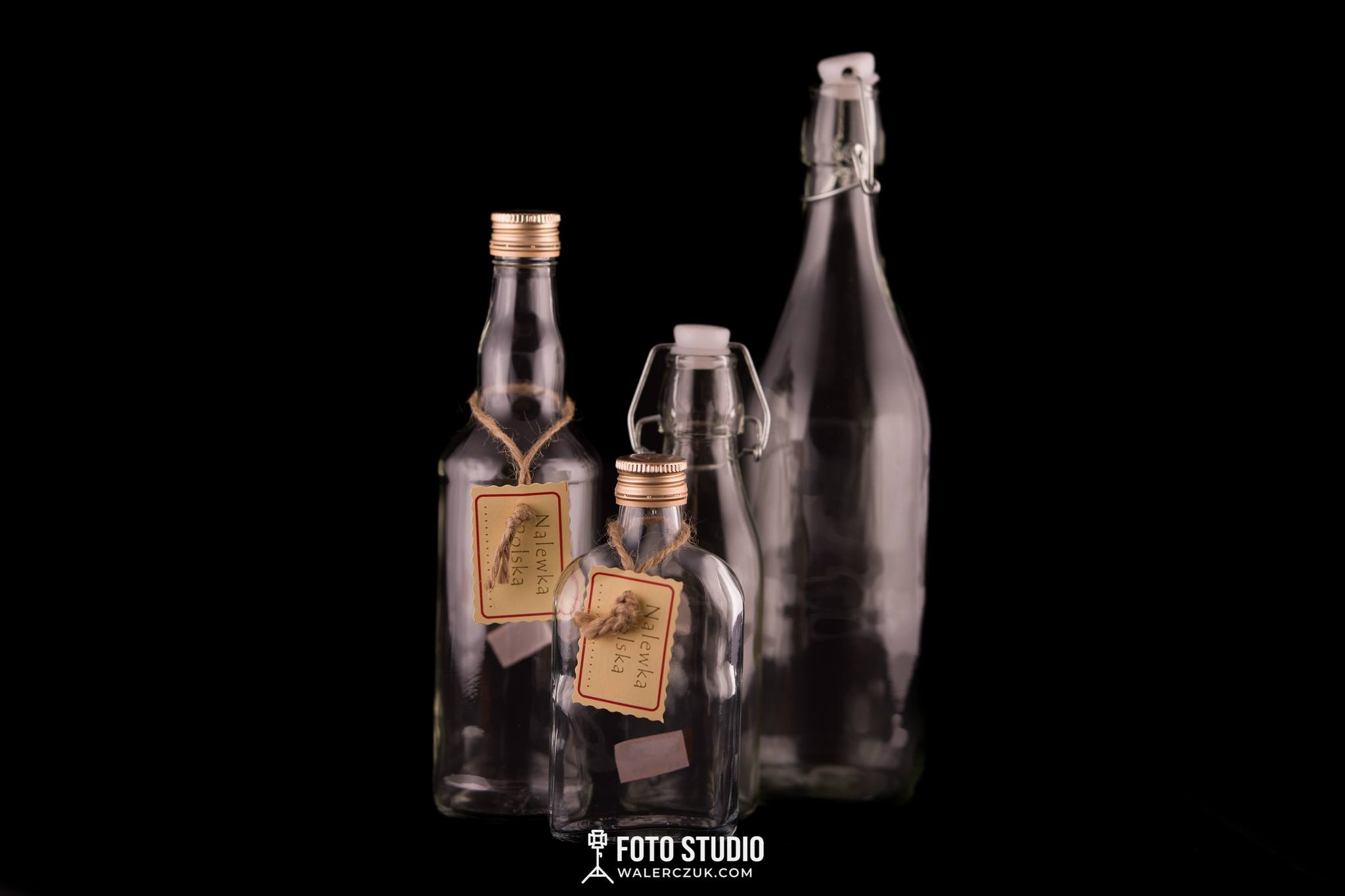 Kompozycja trzech szklanych butelek na czarnym tle, dwie z etykietami 'Nalewka Polska' zawieszonymi na sznurku, oświetlenie studyjne podkreślające kształty.