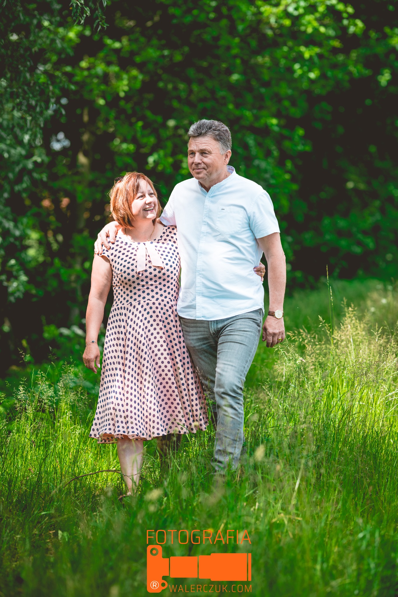 Uśmiechnięta para, kobieta w sukience w grochy i mężczyzna w jasnej koszuli, spacerują objęci pośród zielonej trawy, w tle drzewa.