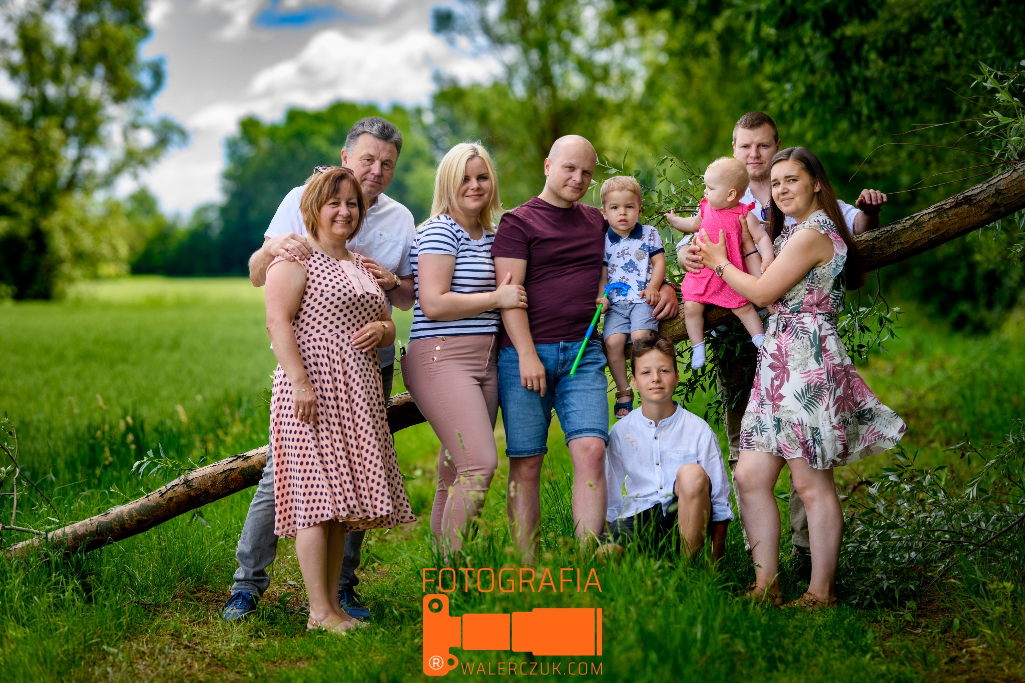 Wielopokoleniowa rodzina pozuje do zdjęcia na tle zielonej łąki i drzew; dzieci siedzą na powalonym pniu drzewa.