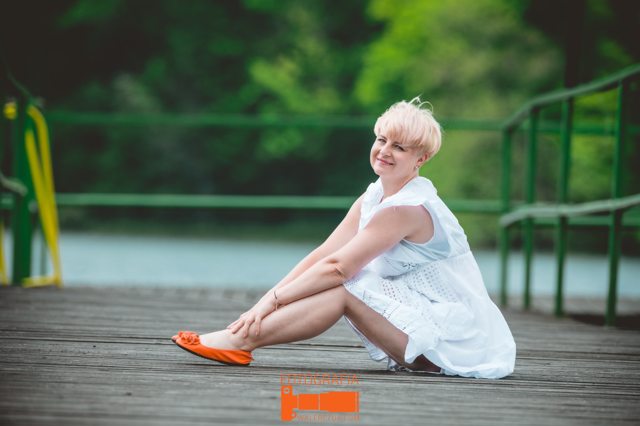 Kobieta z krótkimi blond włosami w białej, ażurowej sukience siedzi na drewnianym pomoście, w tle zielona balustrada i woda, a na stopach ma pomarańczowe baleriny z kwiatkiem.