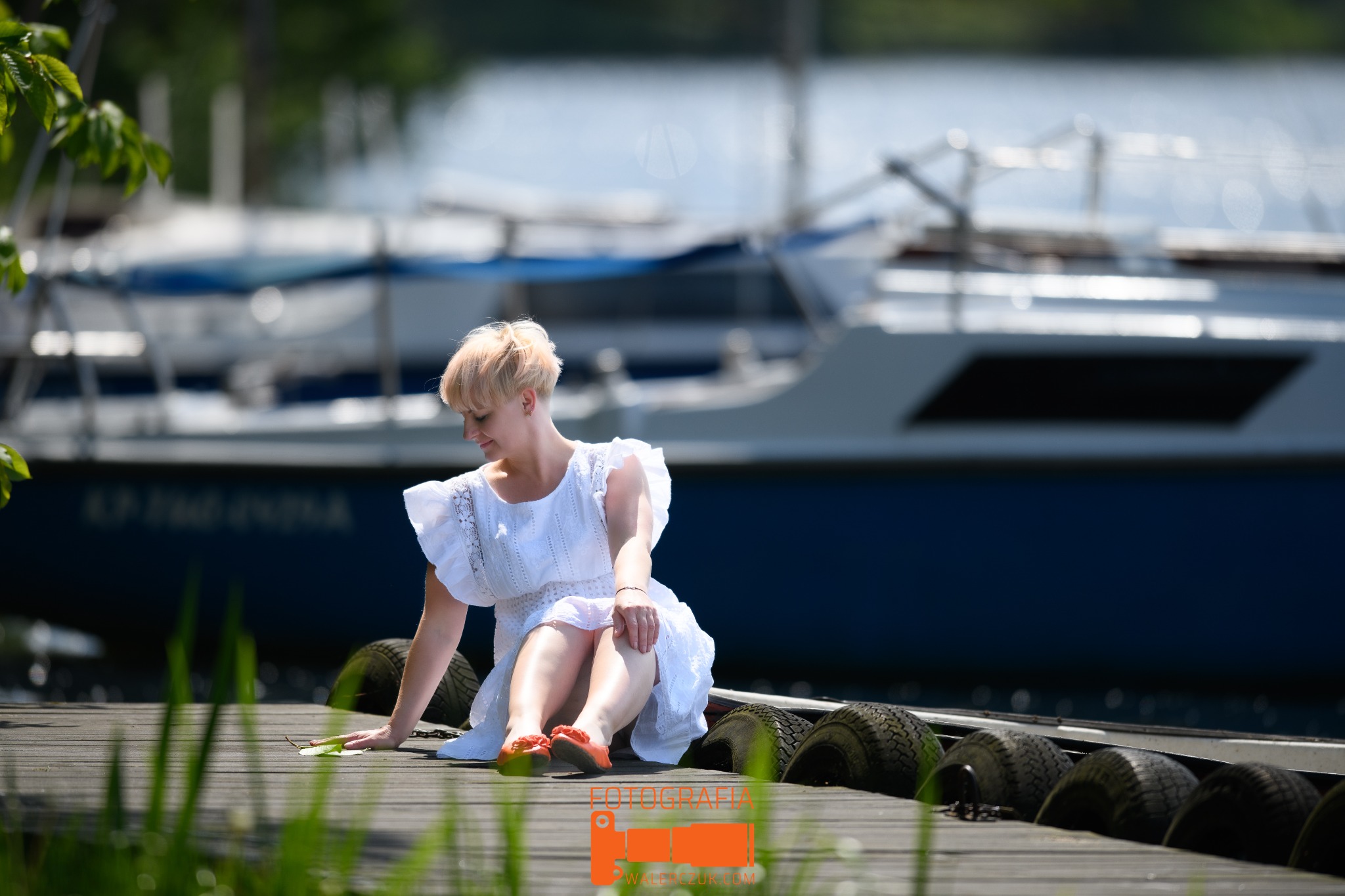 Kobieta w białej sukience siedzi na drewnianym pomoście, w tle zacumowany jacht i woda jeziora.