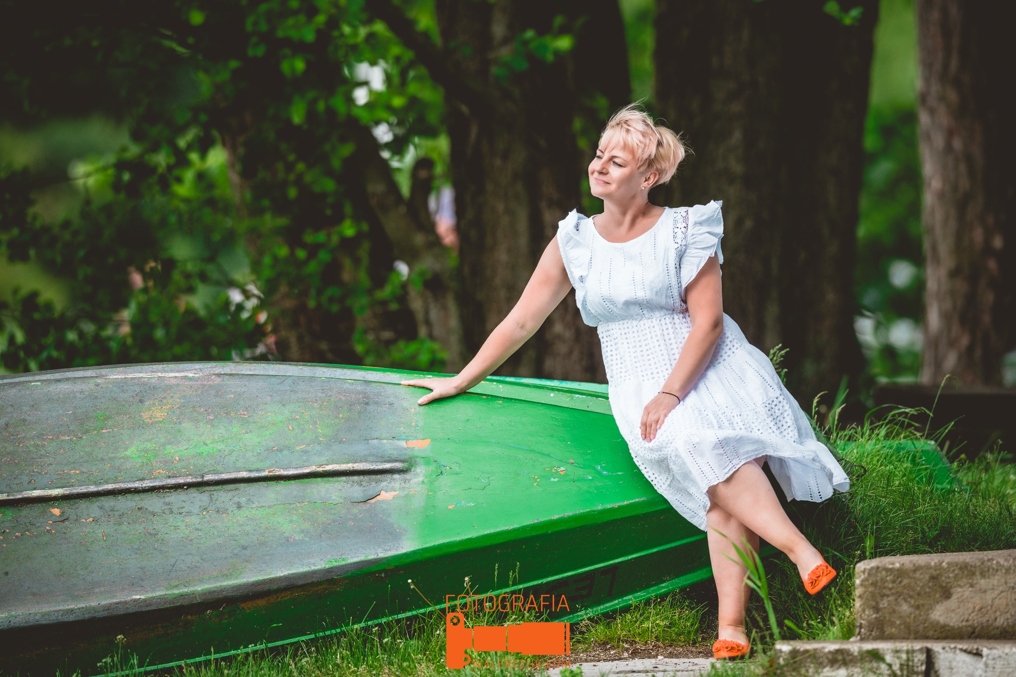 Kobieta w białej sukience opiera się o dnem do góry wywróconą, zieloną łódkę na tle drzew w parku, uśmiechając się w bok.