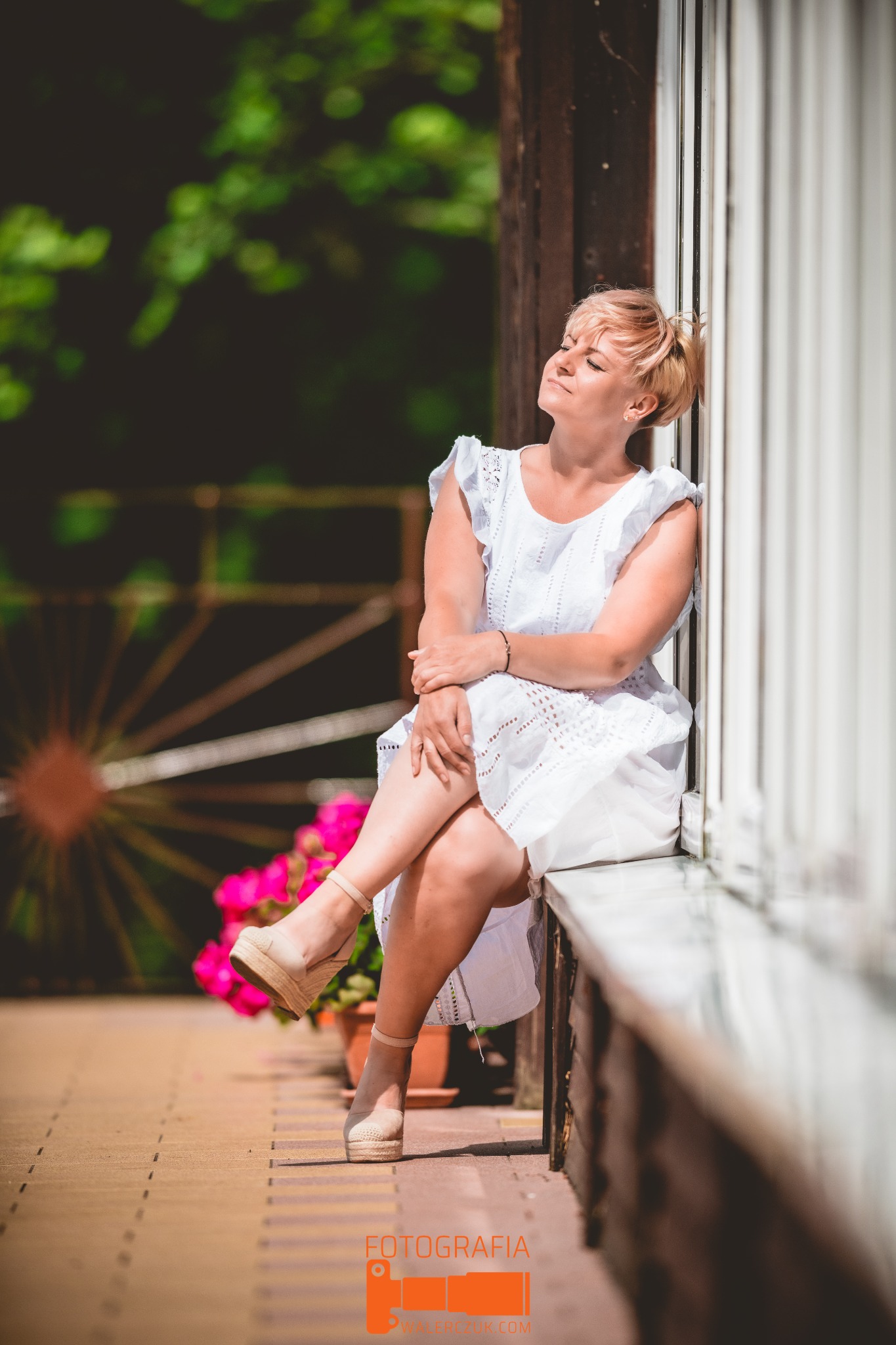 Kobieta w białej, ażurowej sukience siedzi na parapecie, opierając się o białe, pionowe listwy, z zamkniętymi oczami i podniesioną twarzą w stronę słońca, obok doniczki z różowymi kwiatami
