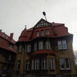 Dwupiętrowa kamienica z czerwonym dachem z dachówki, widoczna z dołu pod pochmurnym niebem, z fragmentem budynku obok z widocznym uszkodzeniem elewacji.