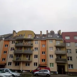 Wielobarwny blok mieszkalny z balkonami i różnymi typami dachów, widok z dołu w pochmurny dzień. Fragment dachu w trakcie naprawy, widoczne uszkodzenia. Na pierwszym planie zaparkowane samochody.