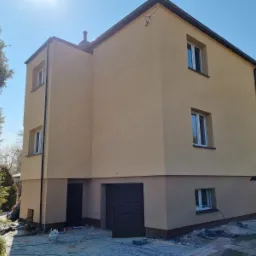 Dwukondygnacyjny dom po tynkowaniu i malowaniu elewacji w odcieniach beżu, z ciemną stolarką okienną i drzwiami garażowymi, widoczny z perspektywy ogrodu w słoneczny dzień.