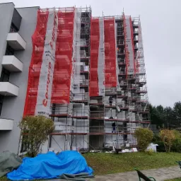 Wielopiętrowy budynek w trakcie prac elewacyjnych, osłonięty rusztowaniami i siatką ochronną z logo producenta farb i tynk&oacute;w, widoczni pracownicy na rusztowaniach, na dole plandeka przykrywająca...