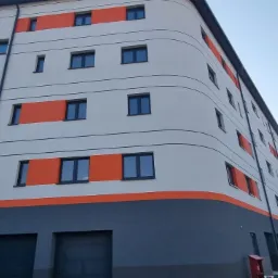 Wielopiętrowy budynek po renowacji elewacji, z szarym dołem i białymi ścianami, pomarańczowe akcenty wokół okien, widoczny pracownik w żółtym kasku.
