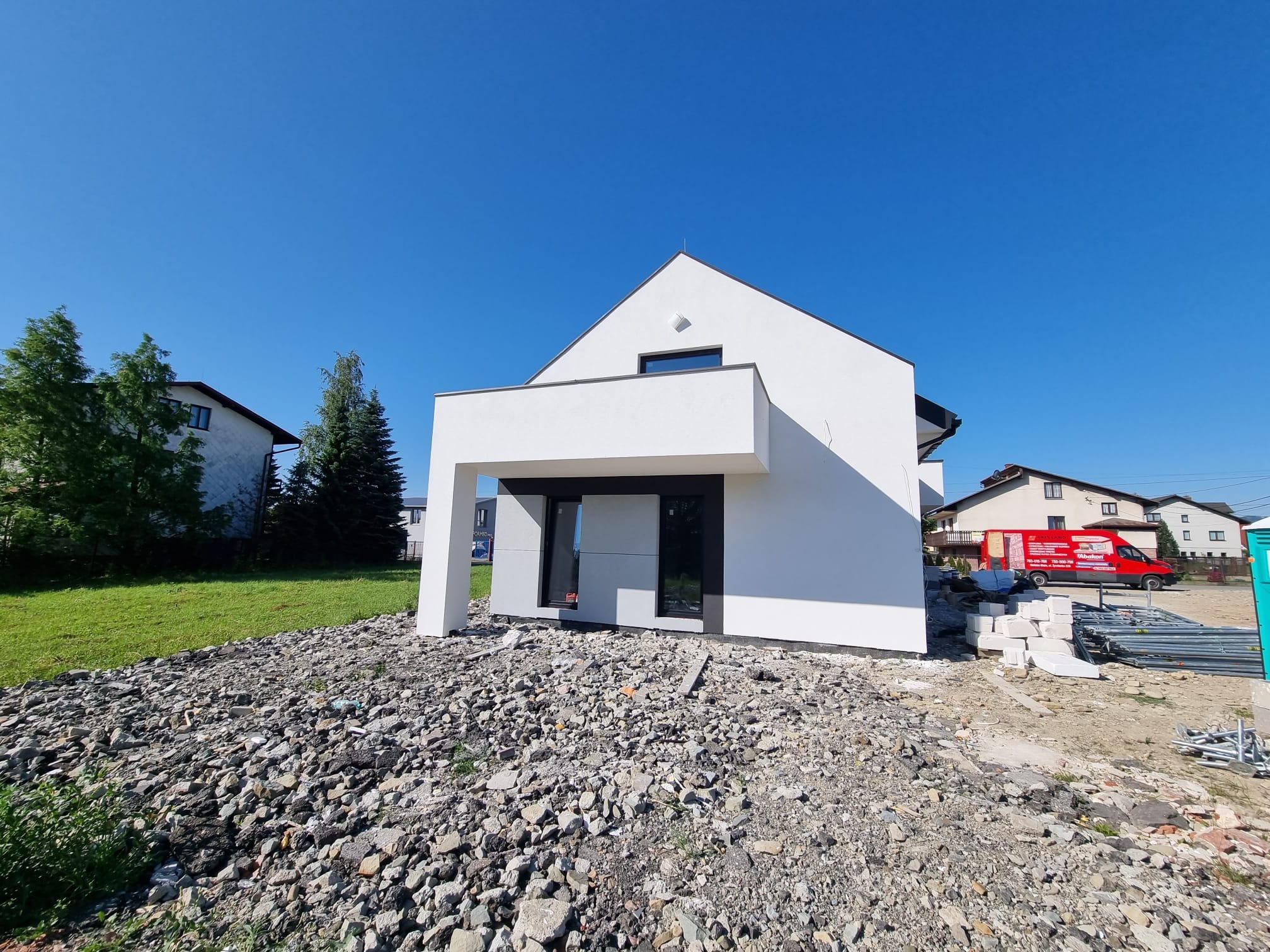 Nowoczesny, biały dom jednorodzinny z płaskim dachem i antracytowymi oknami, widoczny z boku na tle błękitnego nieba. Teren wokół domu częściowo pokryty gruzem, w tle widoczny czerwony samochód...