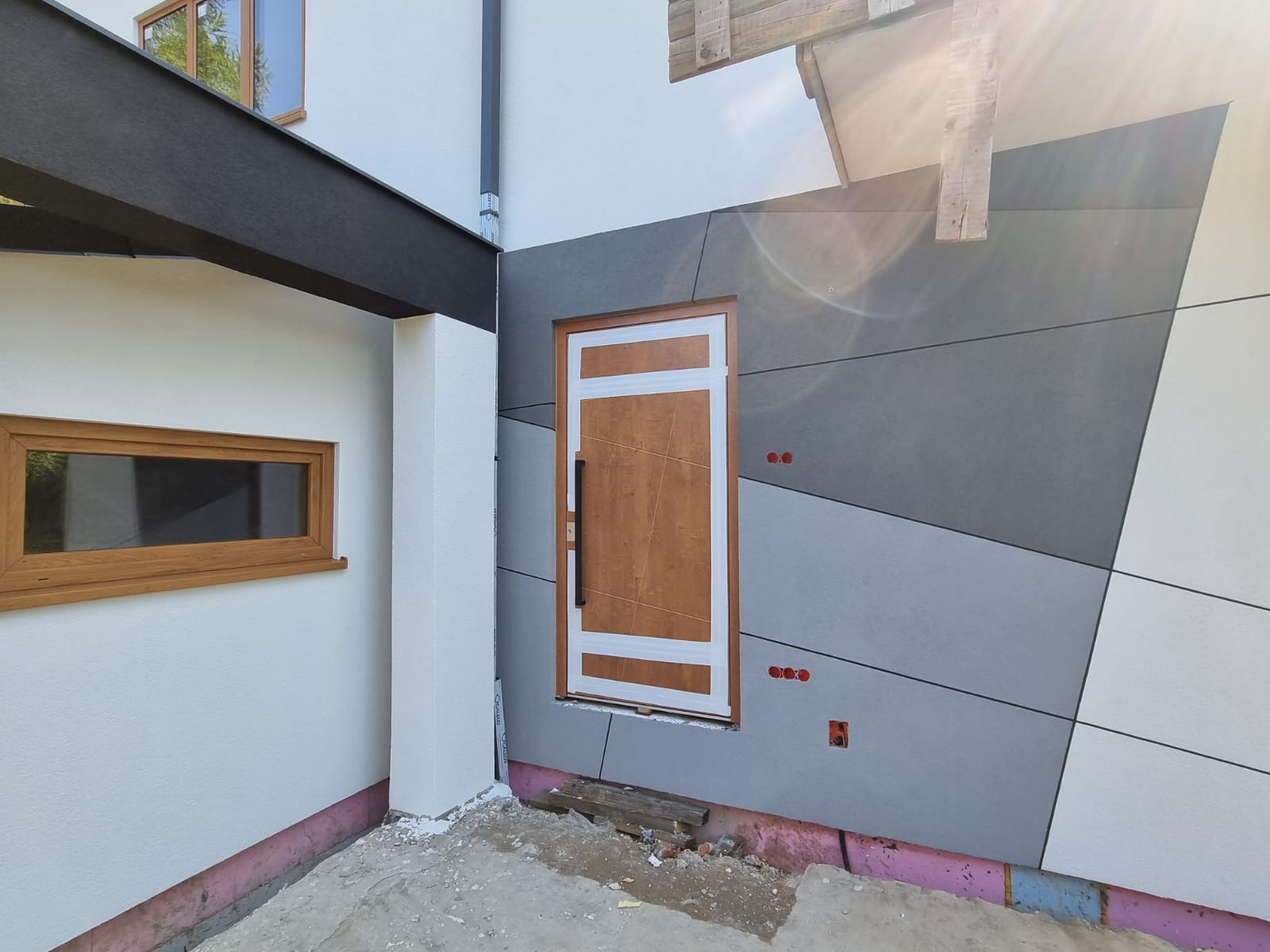 Nowoczesna elewacja domu z drewnianymi oknami i drzwiami, wykończona panelami w odcieniach szarości i bieli, widoczne przygotowania do montażu.