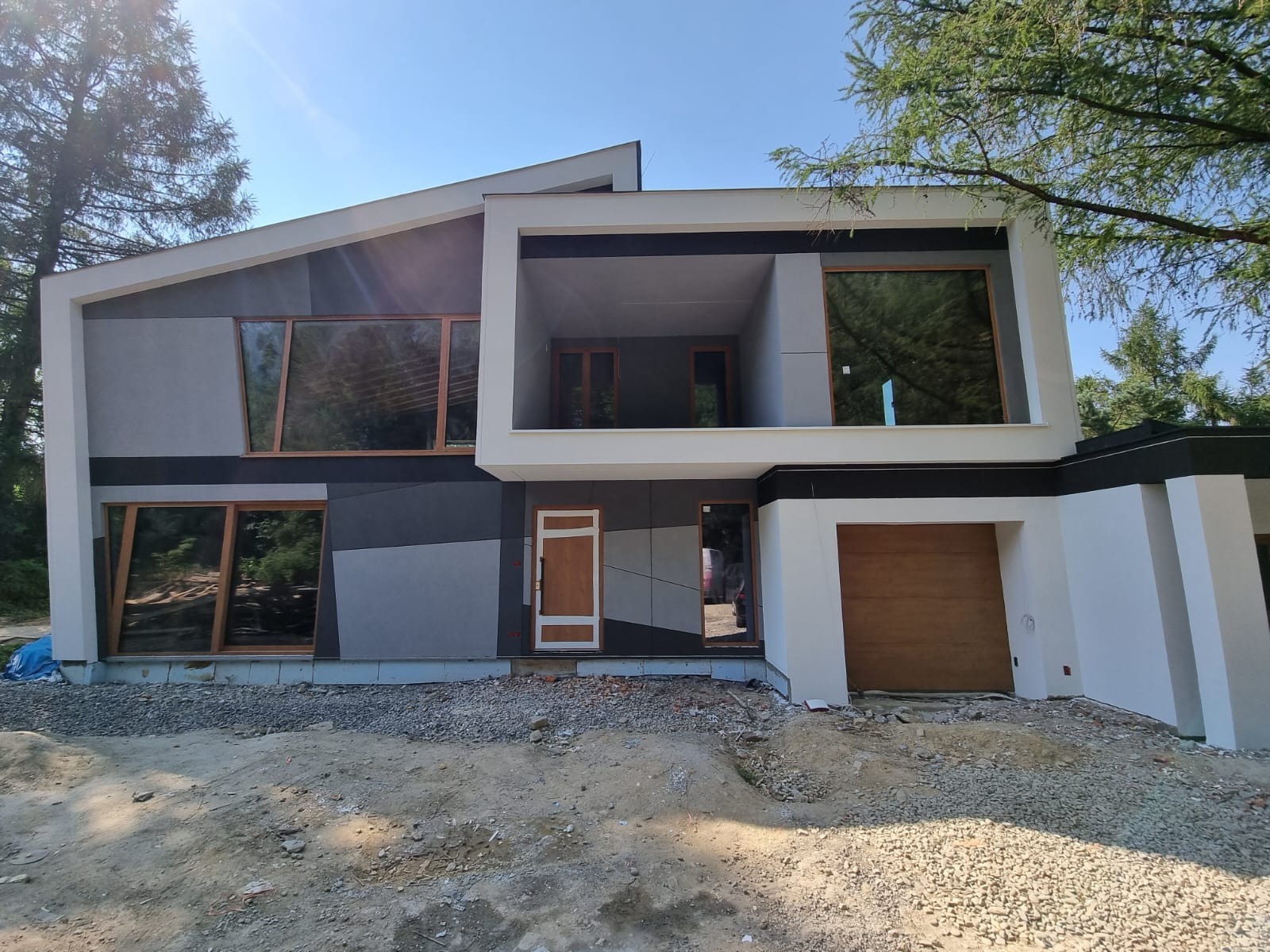Nowoczesny dom z elewacją w odcieniach szarości i bieli, z dużymi oknami o drewnianych ramach, widok na częściowo zagospodarowany teren przed budynkiem.