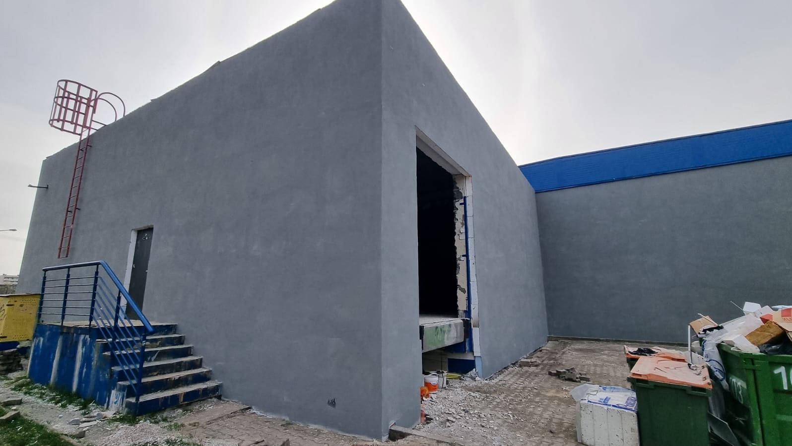 Szary budynek przemysłowy z nową elewacją, widoczne schody z niebieską barierką, drabina z zabezpieczeniem na dachu i otwarty wjazd bez bramy.
