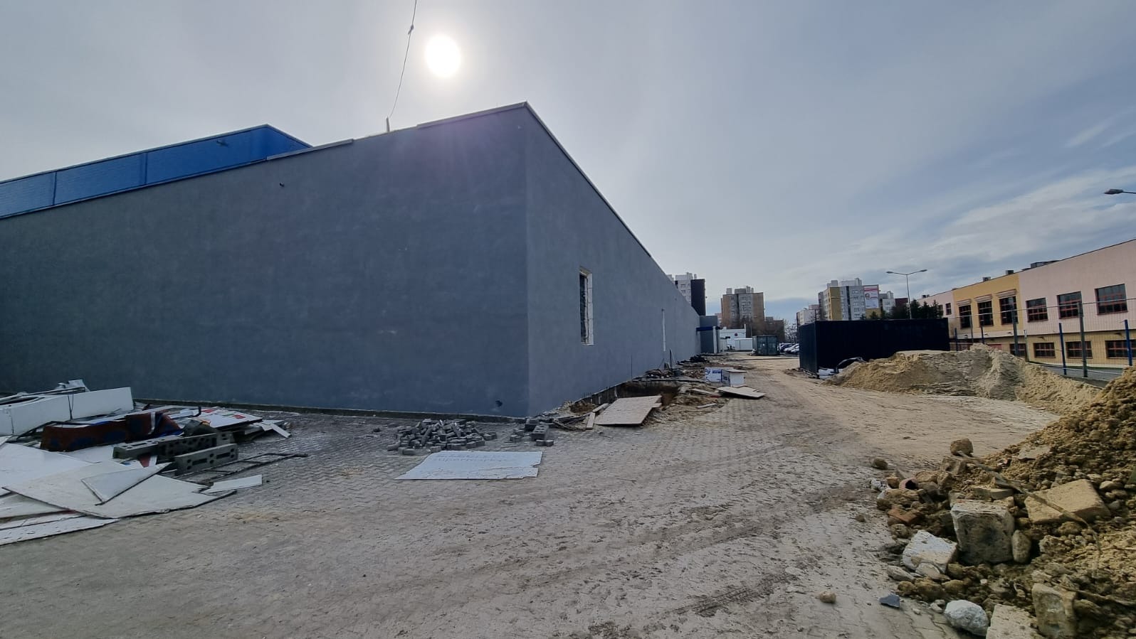 Świeżo otynkowana szara elewacja budynku komercyjnego, widoczny fragment okna bez ramy, teren budowy z gruzem i piaskiem, w tle zabudowa miejska.