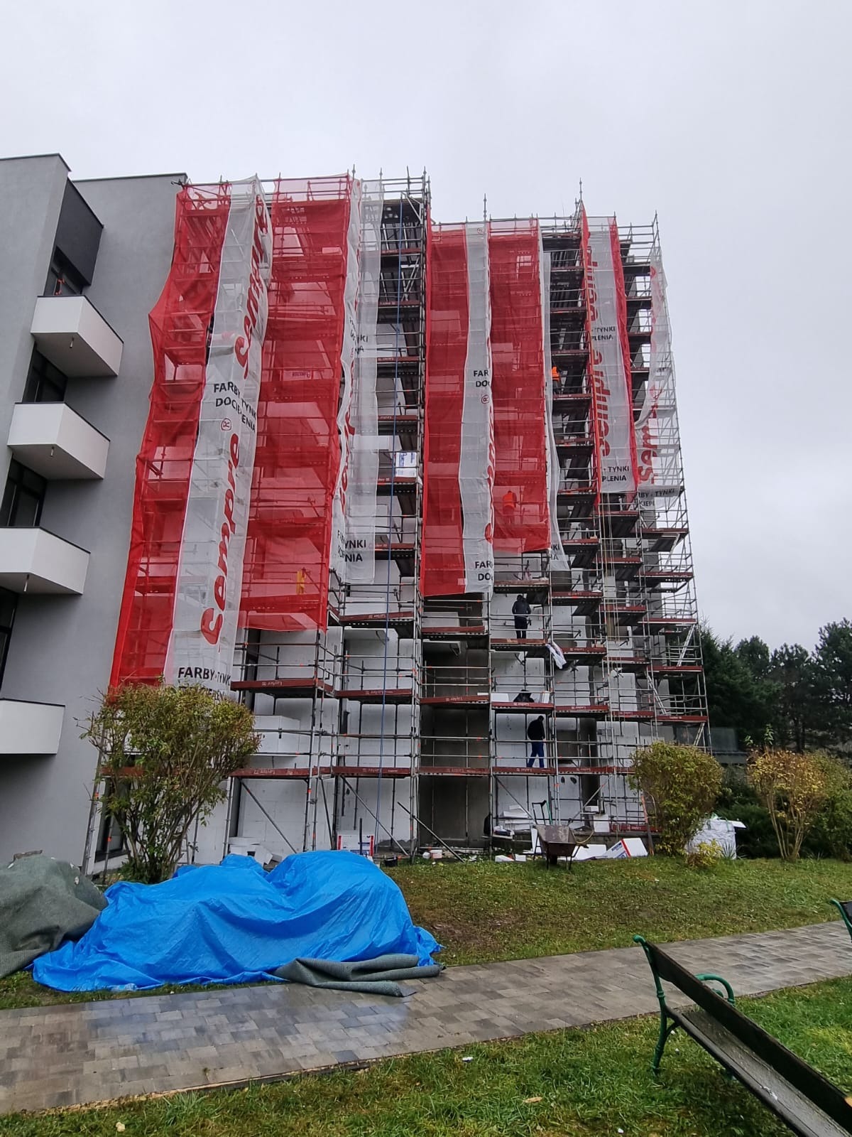 Wielopiętrowy budynek w trakcie prac elewacyjnych, osłonięty rusztowaniami i siatką ochronną z logo producenta farb i tynków, widoczni pracownicy na rusztowaniach, na dole plandeka przykrywająca...