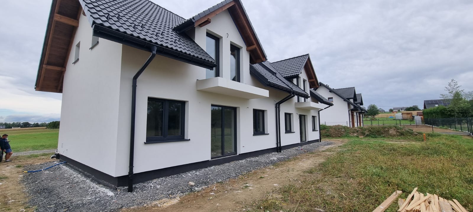 Nowoczesne domy jednorodzinne z białymi elewacjami, czarną stolarką okienną i dachówką, widok z zewnątrz na tle pochmurnego nieba i terenu z gruzem.