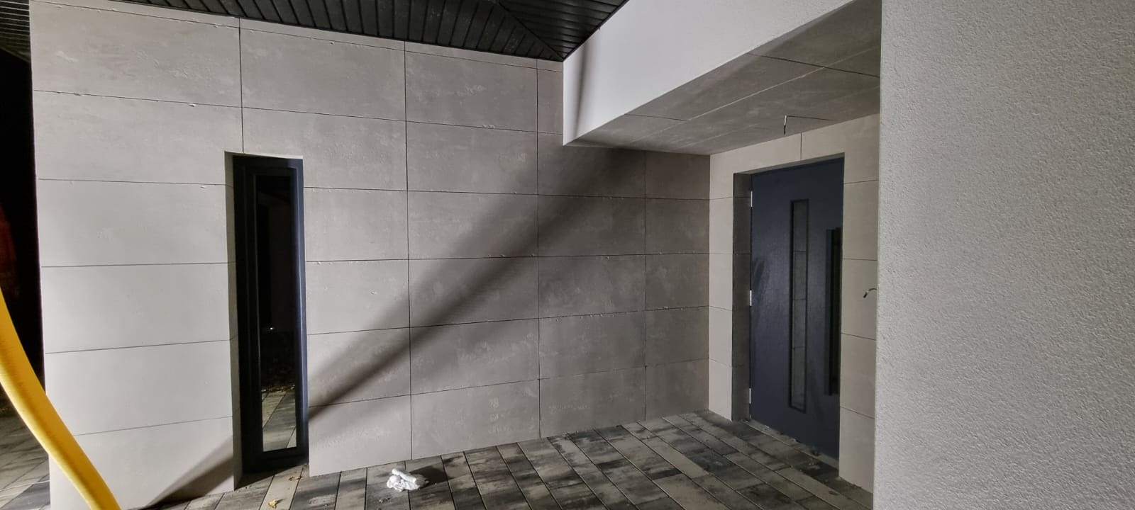 Nowoczesna elewacja budynku z szarych paneli, wąskie okno i ciemne drzwi wejściowe, wykończony chodnik z kostki brukowej.