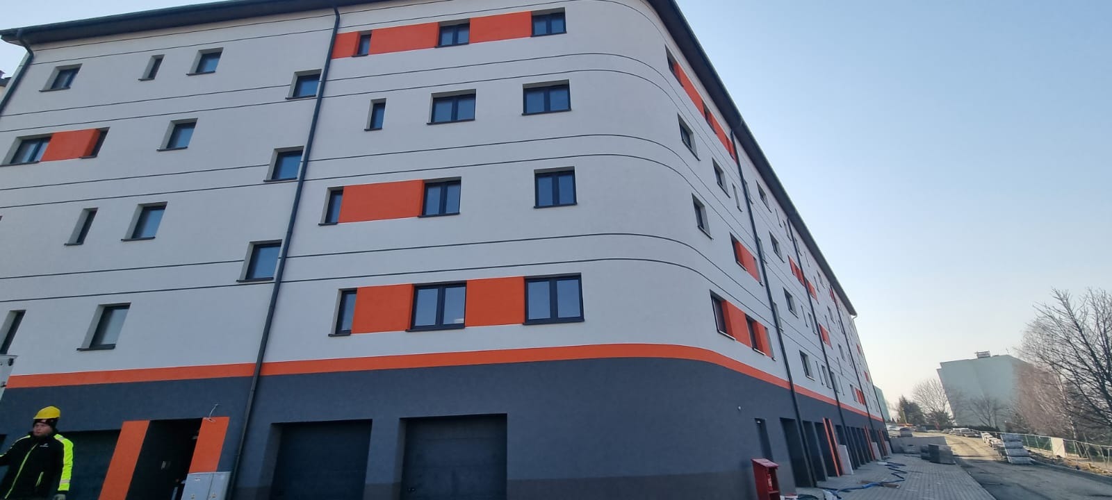 Wielopiętrowy budynek po renowacji elewacji, z szarym dołem i białymi ścianami, pomarańczowe akcenty wokół okien, widoczny pracownik w żółtym kasku.