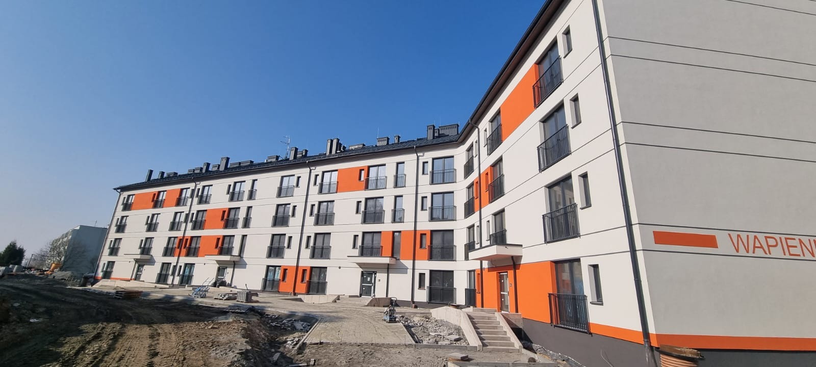 Nowoczesny budynek wielorodzinny po renowacji elewacji, z białymi ścianami i pomarańczowymi akcentami, czarnymi balkonami, widoczne prace brukarskie przed budynkiem.