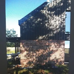 Nowoczesny budynek z ciemną, drewnianą elewacją i ceglanym murem, oświetlony promieniami słońca padającymi przez korony drzew, widoczne zarysy mebli ogrodowych pod zadaszeniem.