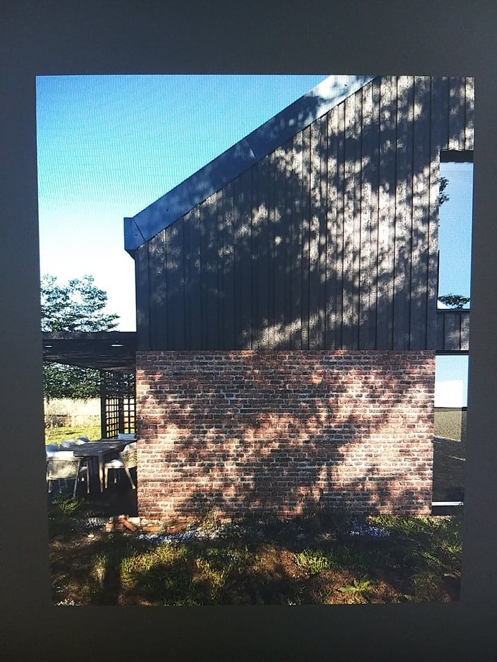 Nowoczesny budynek z ciemną, drewnianą elewacją i ceglanym murem, oświetlony promieniami słońca padającymi przez korony drzew, widoczne zarysy mebli ogrodowych pod zadaszeniem.