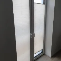 Białe, plisowane osłony okienne w szarym ościeżnicy, częściowo opuszczone, widok z wnętrza pomieszczenia o szarych ścianach i jasnej podłodze.