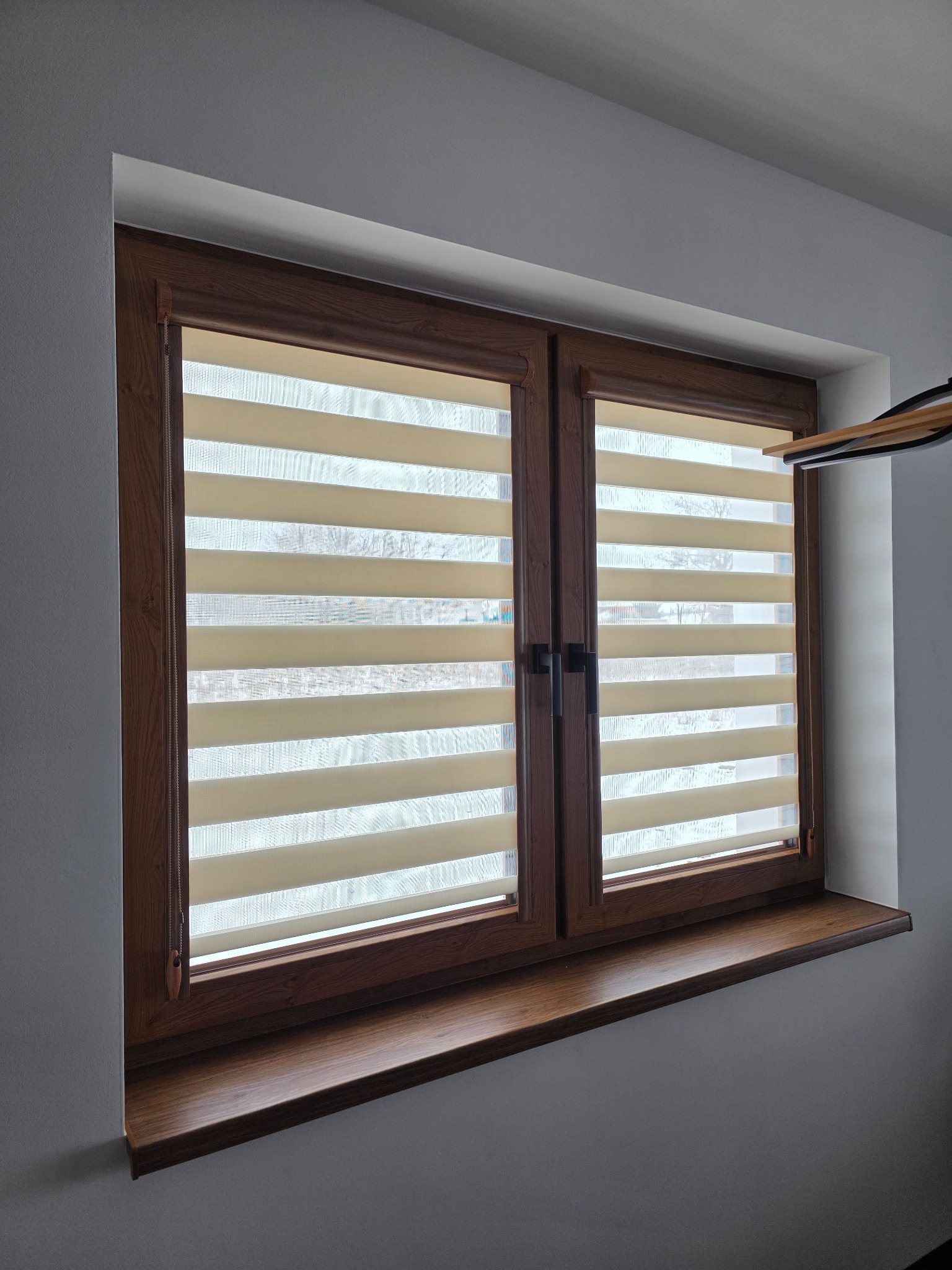 Okno z drewnianą ramą i beżowymi roletami dzień noc w poziome pasy, częściowo zasłonięte, widok na zewnątrz, szare ściany, parapet z drewna.