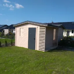 Niewielki, jasny domek narzędziowy z ciemnoszarymi drzwiami i ażurową ścianą, stojący na trawniku przed domami jednorodzinnymi w słoneczny dzień.