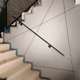 Schody w trakcie wykończenia, zabezpieczone taśmą malarską, z zamontowaną czarną balustradą i dekoracyjną ścianą w odcieniach szarości z geometrycznym wzorem z czarnych linii.