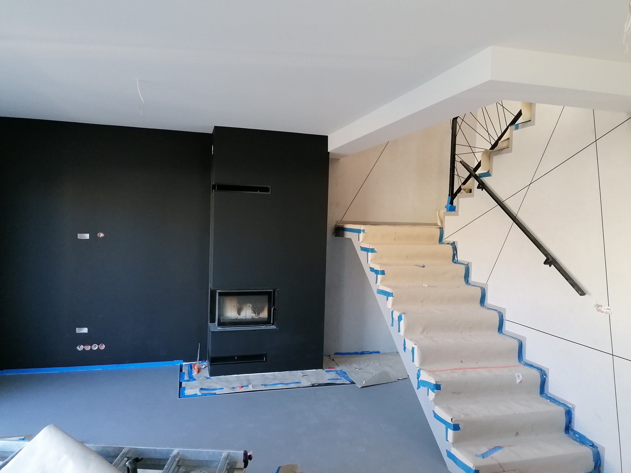 Wnętrze w trakcie remontu: czarna ściana z kominkiem, zabezpieczone schody z metalową balustradą, surowe ściany z geometrycznym wzorem, widoczne puszki elektryczne i taśma malarska.