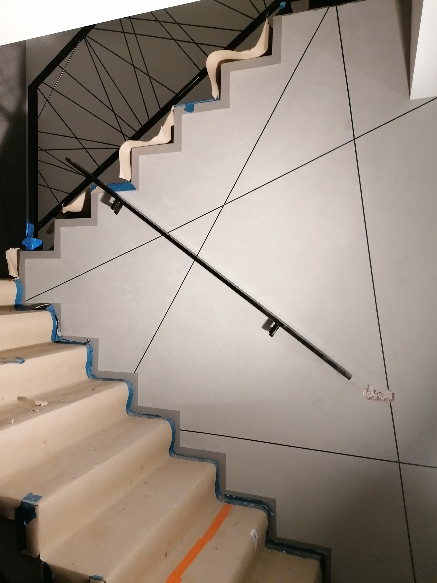 Schody w trakcie wykończenia, zabezpieczone taśmą malarską, z zamontowaną czarną balustradą i dekoracyjną ścianą w odcieniach szarości z geometrycznym wzorem z czarnych linii.