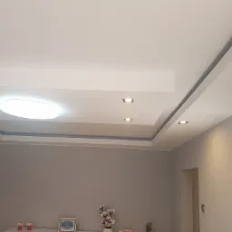 Wykończony sufit podwieszany z oświetleniem punktowym i centralnym okrągłym panelem LED, widok na szarą ścianę i dekoracje na komodzie.