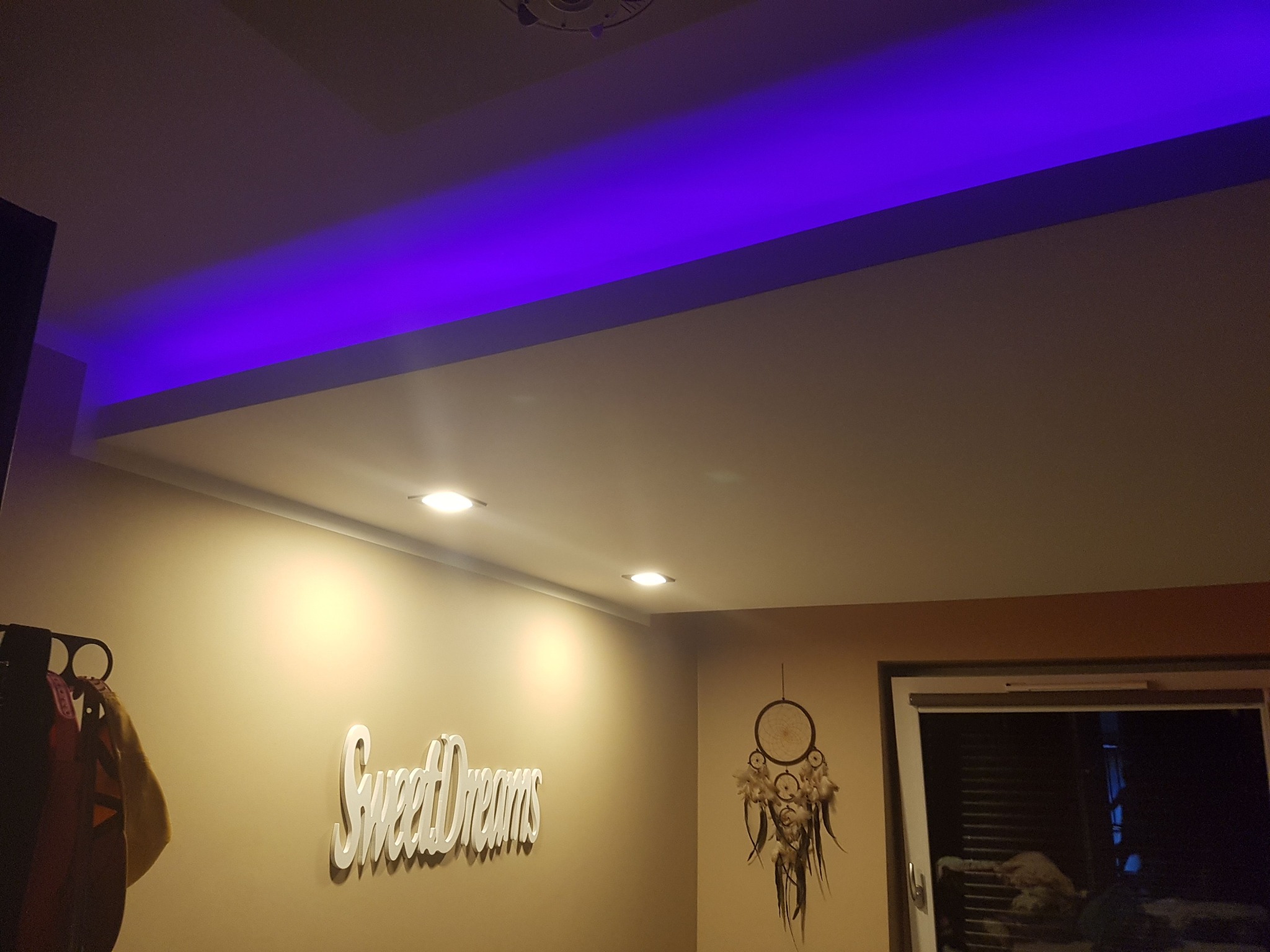Pokój z podwieszonym sufitem z oświetleniem LED w kolorze fioletowym, centralne punkty świetlne, dekoracja na ścianie z napisem 'Sweet Dreams' i łapacz snów.