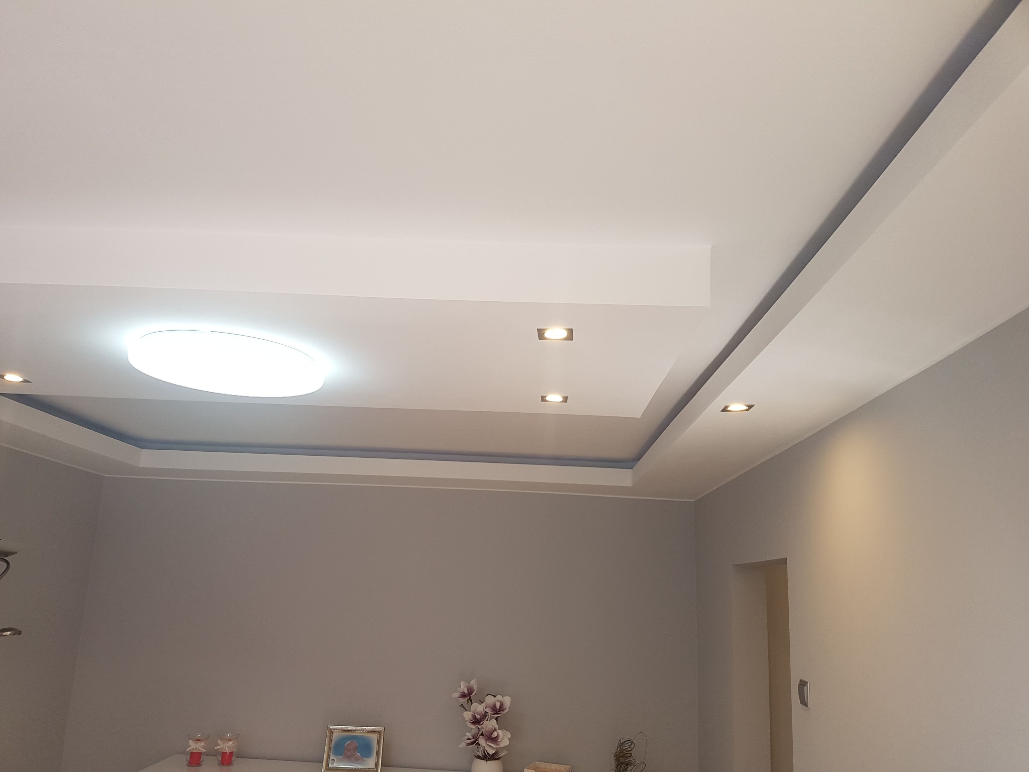 Wykończony sufit podwieszany z oświetleniem punktowym i centralnym okrągłym panelem LED, widok na szarą ścianę i dekoracje na komodzie.